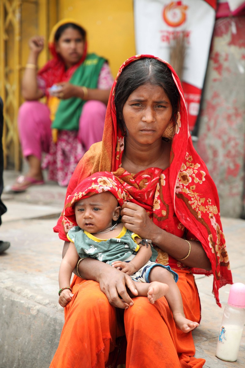 Das Bild zeigt eine Frau in traditioneller Kleidung, die ein Baby auf ihrem Schoß hält, während im Hintergrund weitere Menschen zu sehen sind.