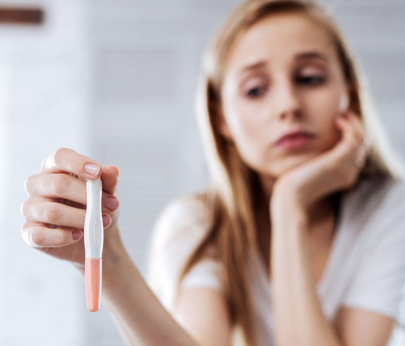 Junge Frau hält einen Schwangerschaftstest in ihren Händen und ihr Blick ist traurig.